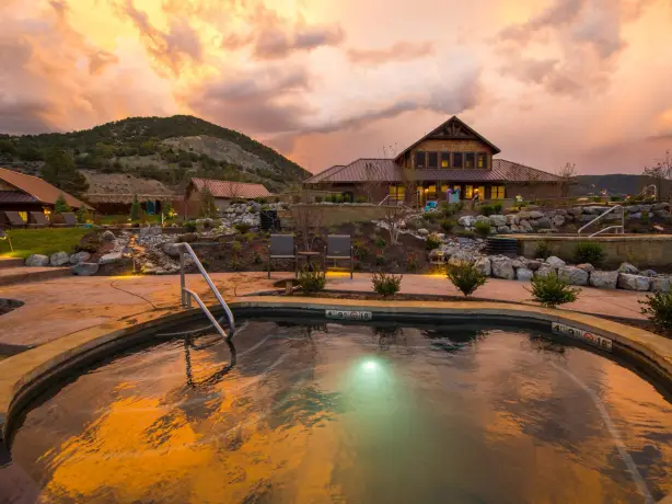 Thermalbecken der Iron Mountain Hot Springs in Glenwood Springs, Colorado, bei Sonnenuntergang. Im Vordergrund spiegelt sich der farbenfrohe Himmel im warmen Wasser, dahinter liegen das Spa-Gebäude, gepflegte Gartenanlagen und die umliegenden Hügel.