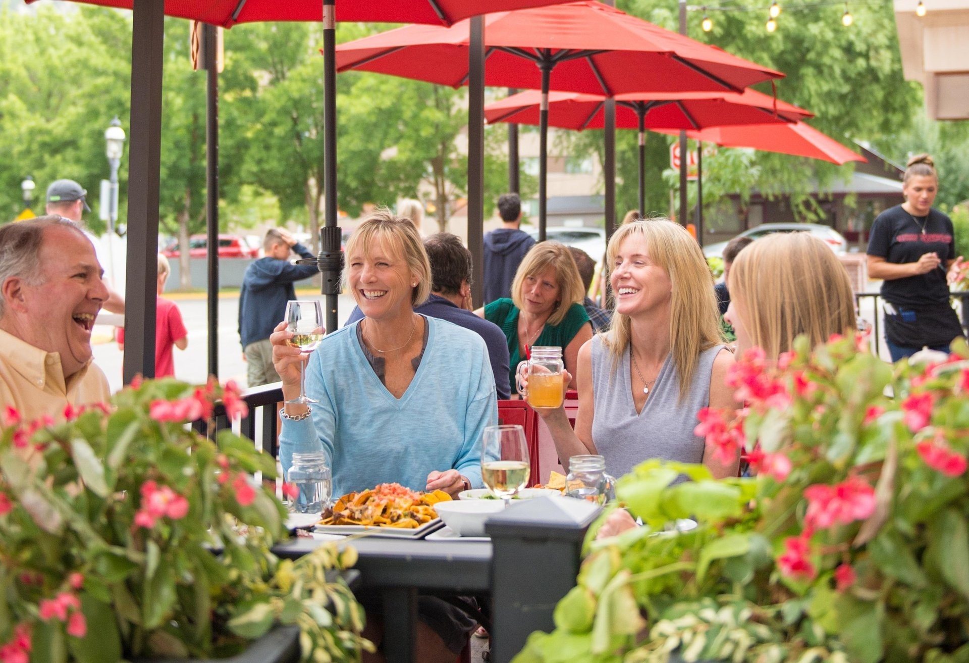 Menschen genießen gemeinsam ein Essen auf der Außenterrasse eines Restaurants in Glenwood Springs, Colorado. Unter roten Sonnenschirmen sitzen sie in entspannter Atmosphäre zwischen blühenden Blumen und stoßen mit Getränken an.