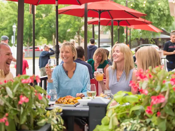 Menschen genießen gemeinsam ein Essen auf der Außenterrasse eines Restaurants in Glenwood Springs, Colorado. Unter roten Sonnenschirmen sitzen sie in entspannter Atmosphäre zwischen blühenden Blumen und stoßen mit Getränken an.