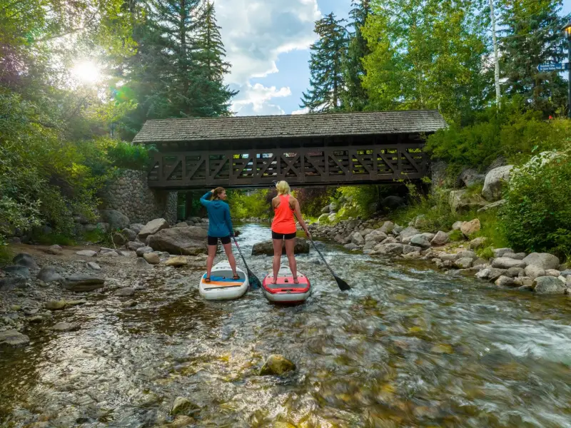 Zwei Stand-up-Paddlerinnen gleiten auf einem flachen Gebirgsbach in Vail, Colorado, unter einer überdachten Holzbrücke hindurch. Umgeben von Felsen, dichtem Grün und hohen Nadelbäumen spiegelt sich das Sonnenlicht im klaren Wasser.