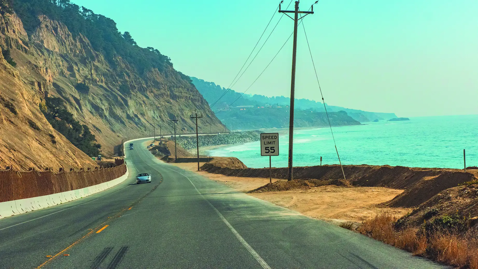 A coastal highway in California curving along steep cliffs above the ocean, with cars driving beside the turquoise water.
