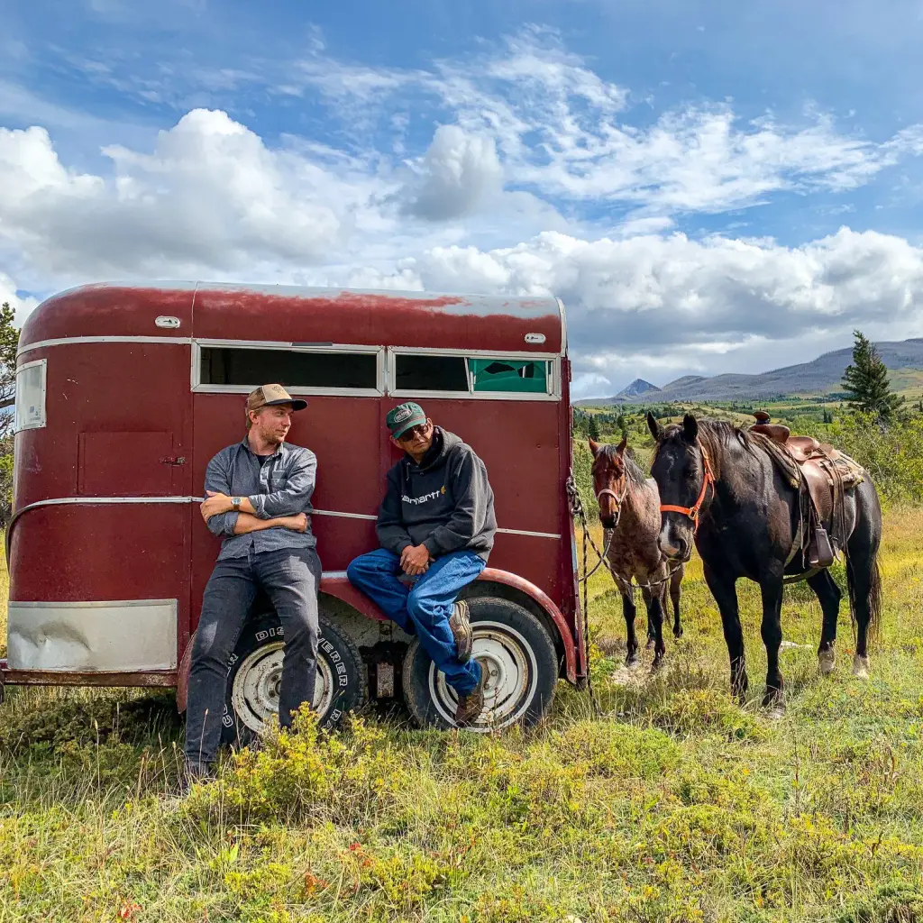 Zwei Männer lehnen an einem roten Anhänger, an dem zwei Pferde angebunden sind, vor dem Hintergrund sanfter Hügel und blauem Himmel in Montana