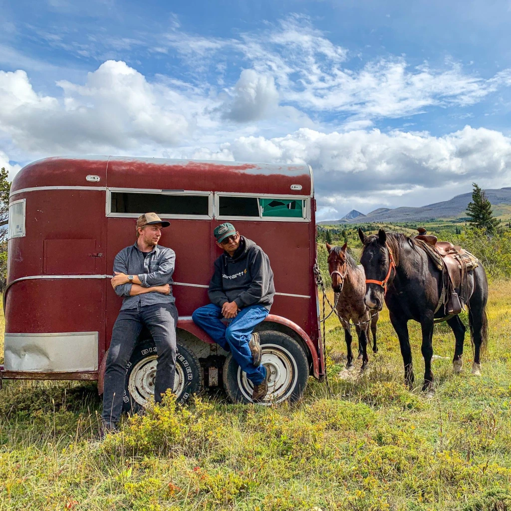 Zwei Männer lehnen an einem roten Anhänger, an dem zwei Pferde angebunden sind, vor dem Hintergrund sanfter Hügel und blauem Himmel in Montana