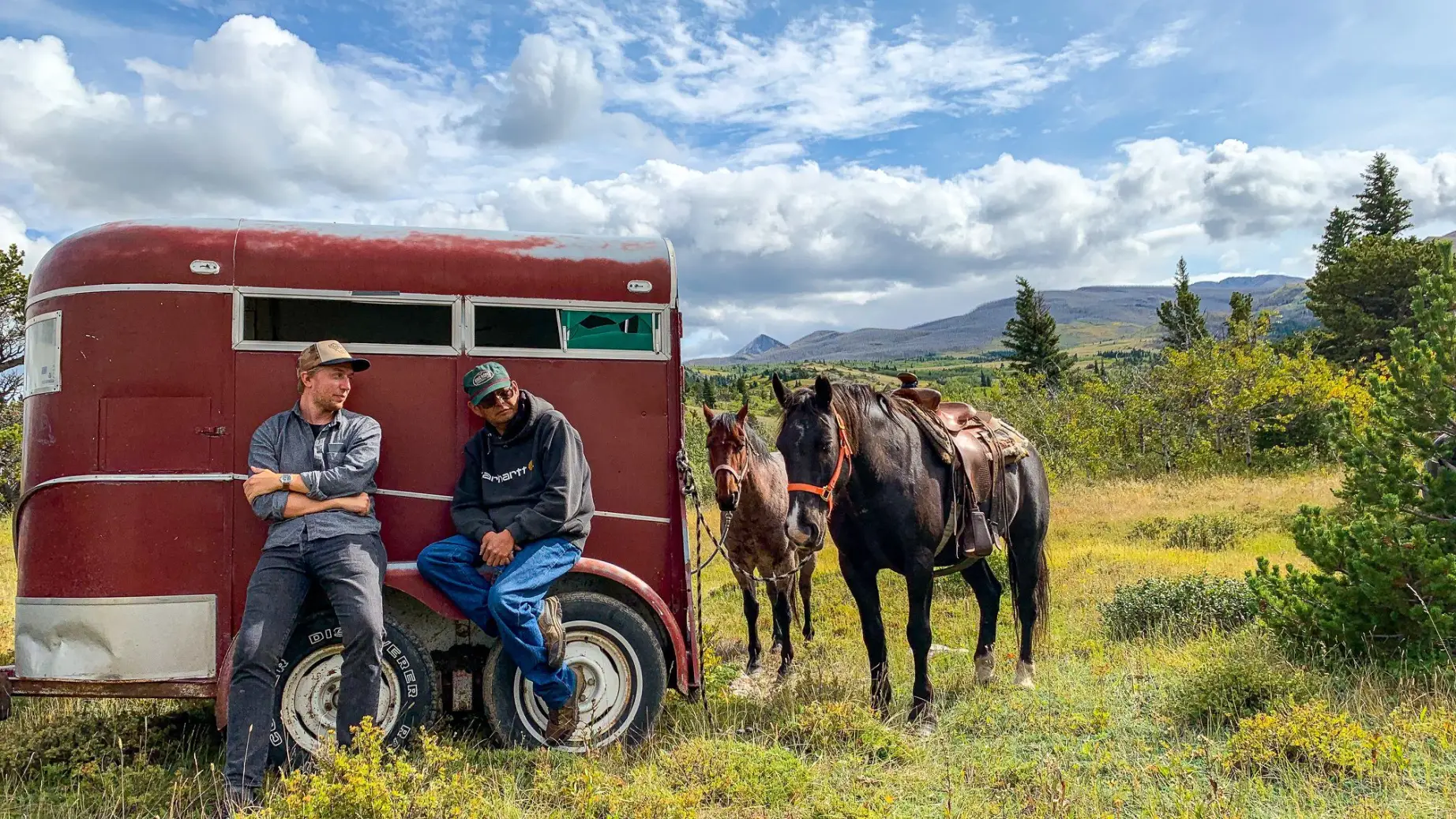 Two men lean against a red trailer which has two horses tied to it, with a background of rolling hills and blue skies in Montana