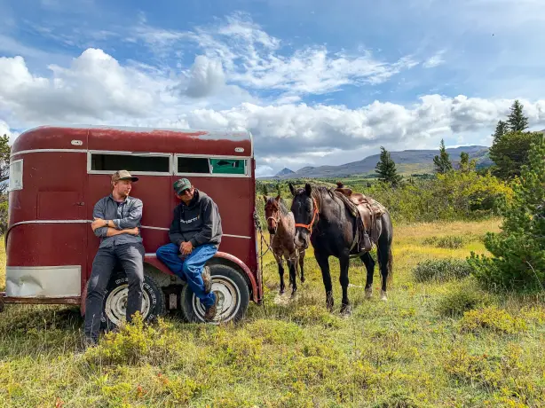 Two men lean against a red trailer which has two horses tied to it, with a background of rolling hills and blue skies in Montana