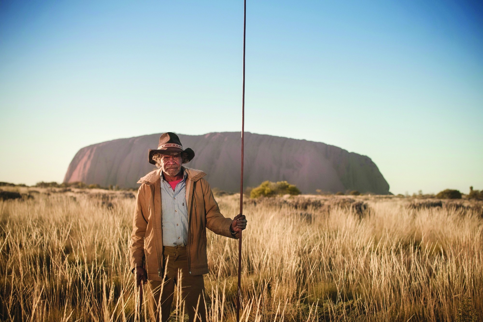 Ein einheimischer Fremdenführer im australischen Uluru-Nationalpark, der im hohen Gras steht und einen Stab in der Hand hält, mit der Felsformation Uluru im Hintergrund.