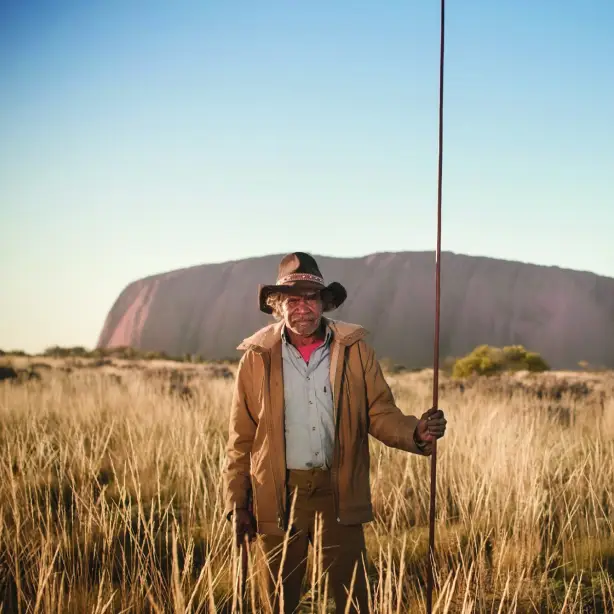 Ein einheimischer Fremdenführer im australischen Uluru-Nationalpark, der im hohen Gras steht und einen Stab in der Hand hält, mit der Felsformation Uluru im Hintergrund.