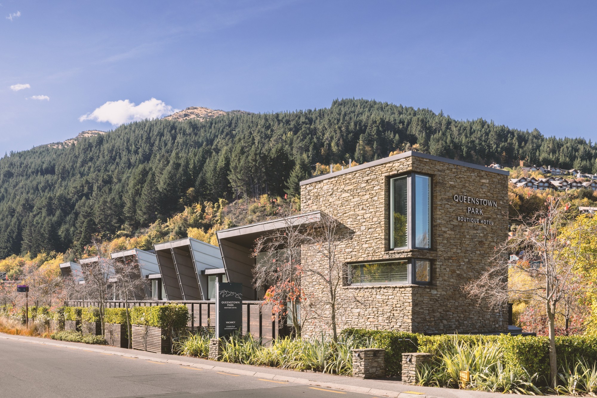 Ein modernes, mit Stein verkleidetes Boutique-Hotel mit kantigen Dachlinien, umgeben von herbstlichem Laub und mit Kiefern bewachsenen Hügeln in Queenstown, Neuseeland