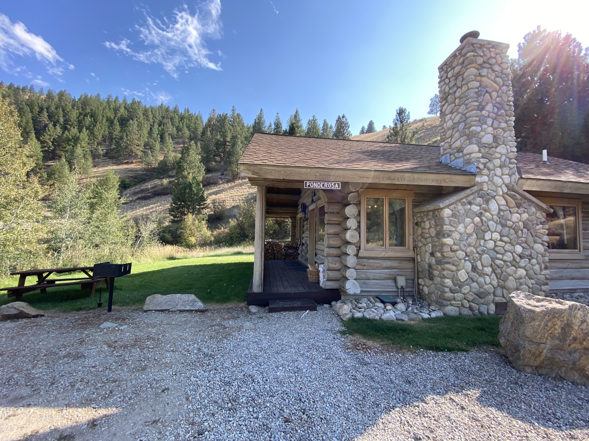 Buitenkant van de Ponderosa blokhut bij Rye Creek Lodge, met een stenen schoorsteen, overdekt terras met gestapeld brandhout en een picknicktafel met BBQ op een grasveld, omgeven door met dennenbomen begroeide heuvels in de Bitterroot Valley, Montana, VS.