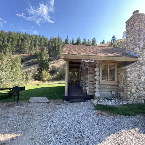 Außenansicht der Ponderosa-Blockhütte in der Rye Creek Lodge, mit einem Steinkamin, überdachter Veranda mit gestapeltem Brennholz und einem Picknicktisch mit Grill auf einer Rasenfläche, eingebettet in die kiefernbewachsenen Hügel des Bitterroot Valley, Montana, USA.