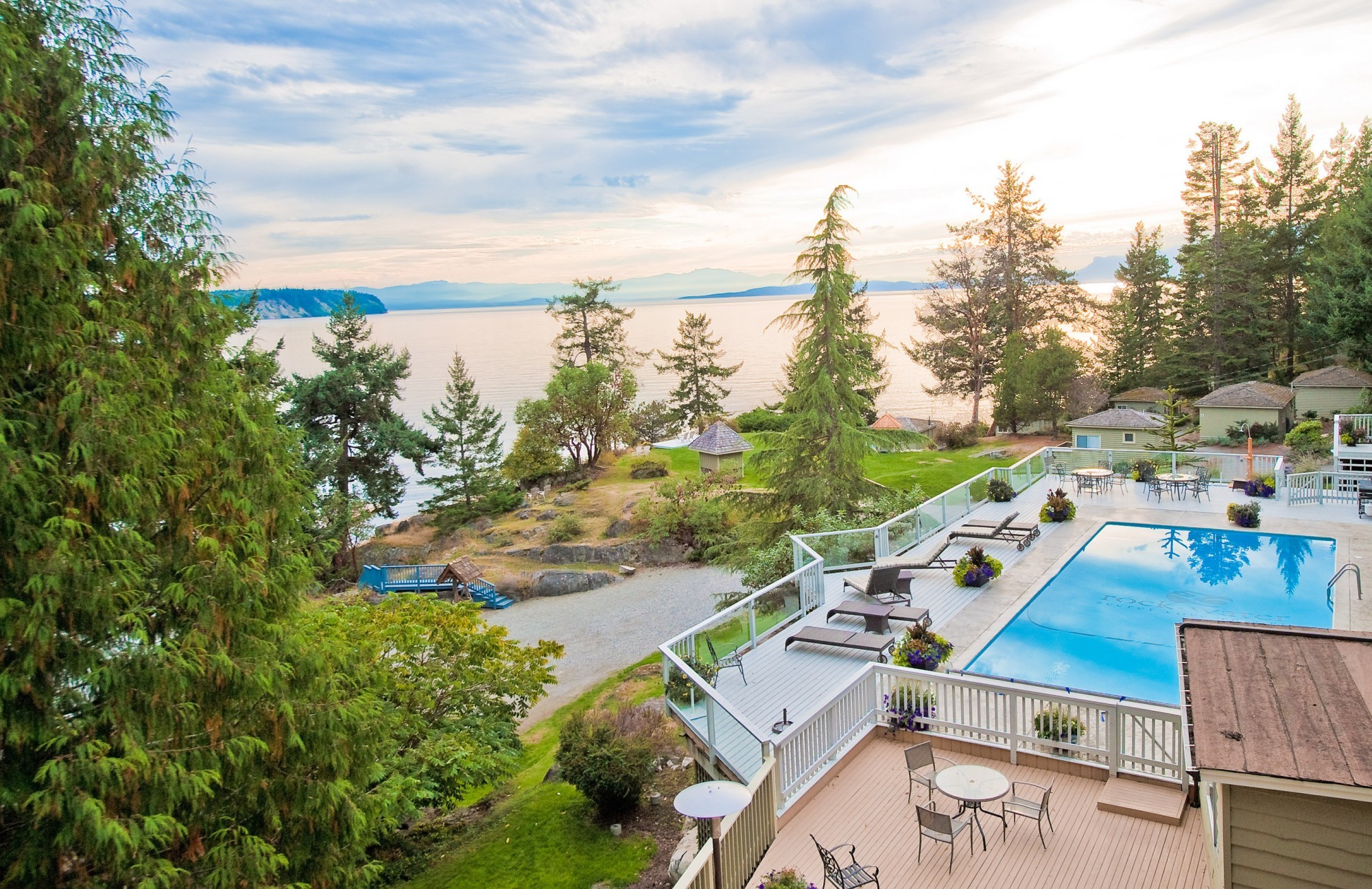 Erhöhter Blick auf den Außenpool und die Sonnenterrasse eines Resorts mit Liegestühlen, mit Blick auf einen angelegten Garten und eine felsige Küstenlinie, ruhiges Meerwasser und ferne Berge unter einem sanften Abendhimmel an der Sunshine Coast, British Columbia, Kanada.