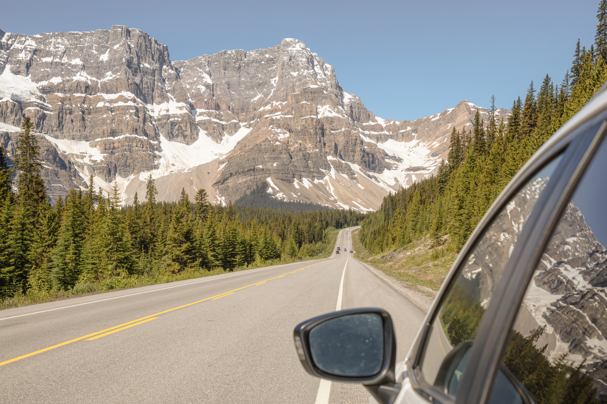 Blick aus einem Autofenster auf dem Icefields Parkway, mit einer geraden Straße, die sich durch Nadelwald auf dramatische schneebedeckte Felsgipfel unter strahlend blauem Himmel zustreckt, in den Kanadischen Rockies, Alberta, Kanada.