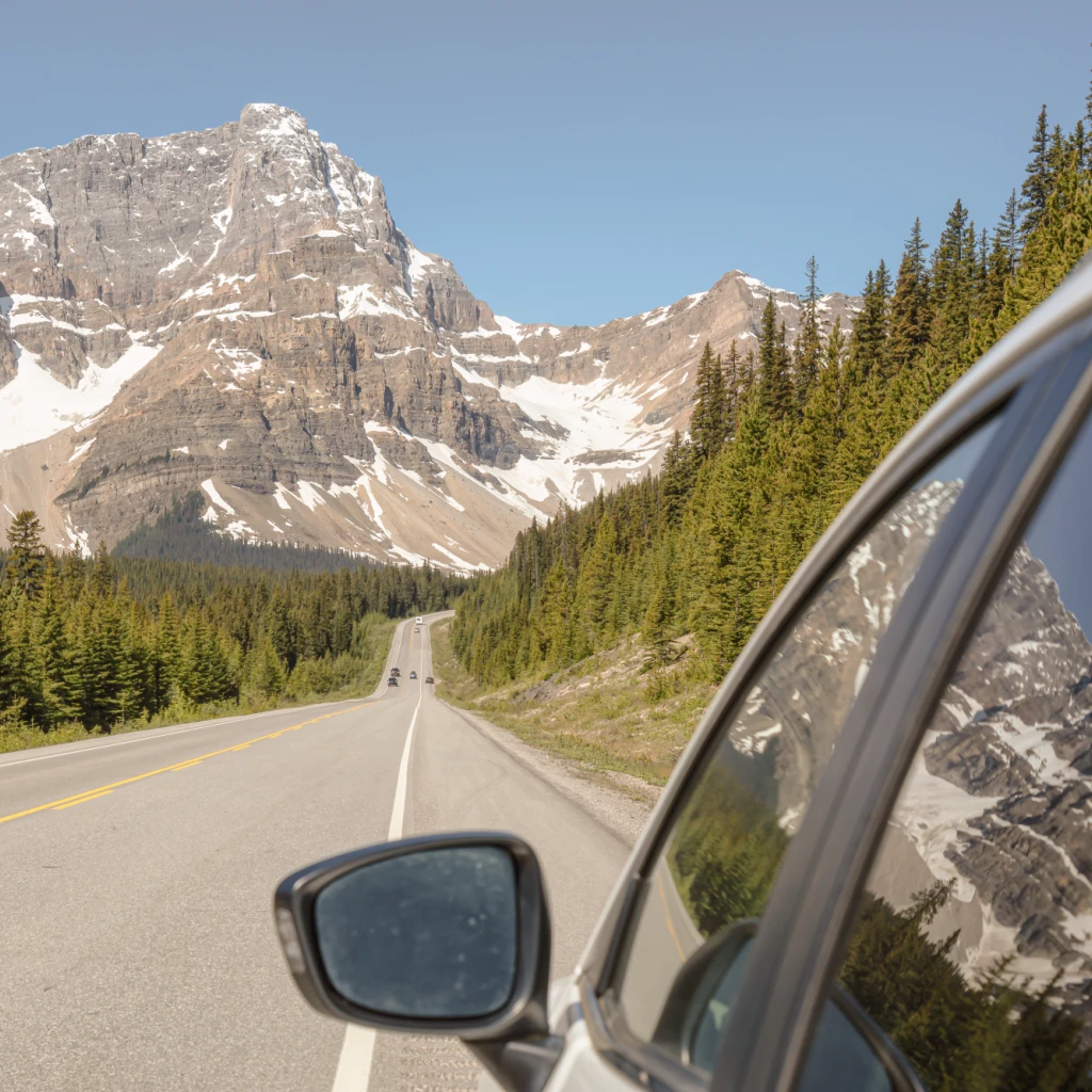 Blick aus einem Autofenster auf dem Icefields Parkway, mit einer geraden Straße, die sich durch Nadelwald auf dramatische schneebedeckte Felsgipfel unter strahlend blauem Himmel zustreckt, in den Kanadischen Rockies, Alberta, Kanada.
