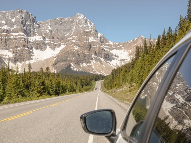View from a car window driving along the Icefields Parkway, with a straight road stretching ahead through pine forest towards dramatic snow-capped rocky peaks under a clear blue sky, in the Canadian Rockies, Alberta, Canada.