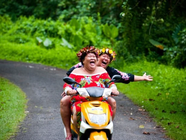 Two Cook Islanders laugh while riding a scooter through the forest