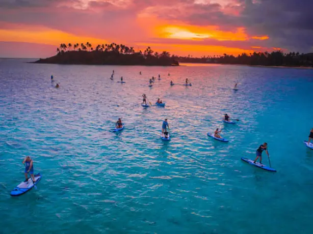 A group of people paddle boarding in the bays of Cook Islands with a colourful sky in the background