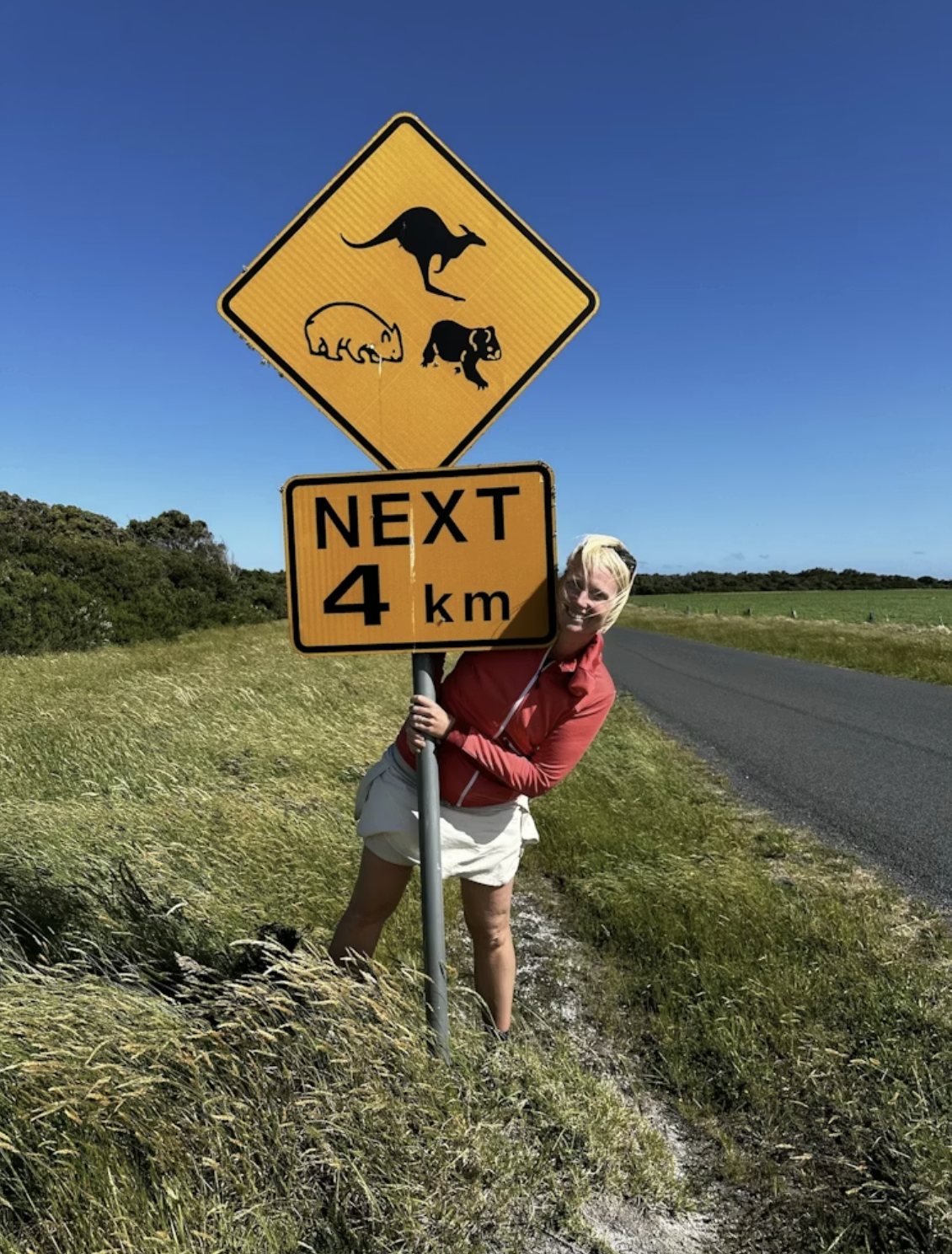 Frau steht neben einer Straße und umfasst ein australisches Verkehrsschild mit Tiersymbolen und Hinweis "Next 4 km"