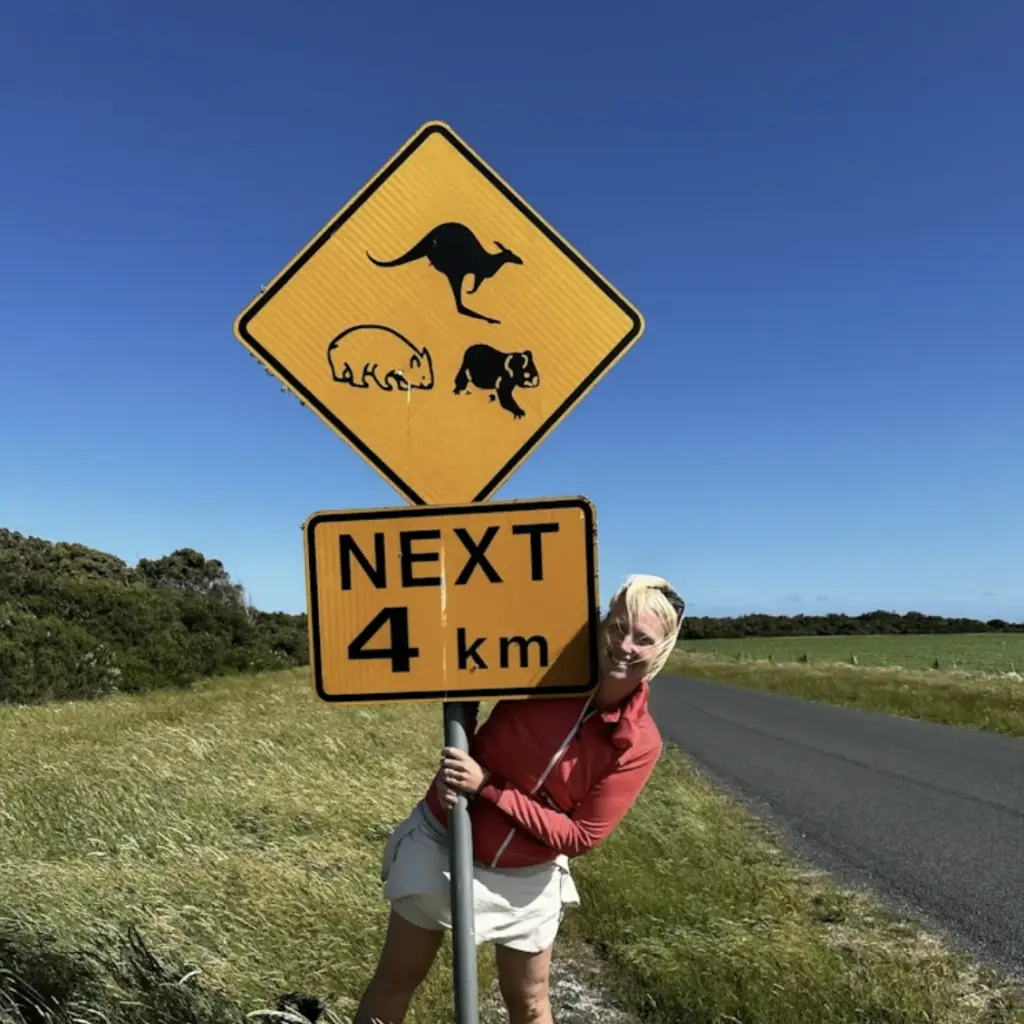Frau steht neben einer Straße und umfasst ein australisches Verkehrsschild mit Tiersymbolen und Hinweis "Next 4 km"