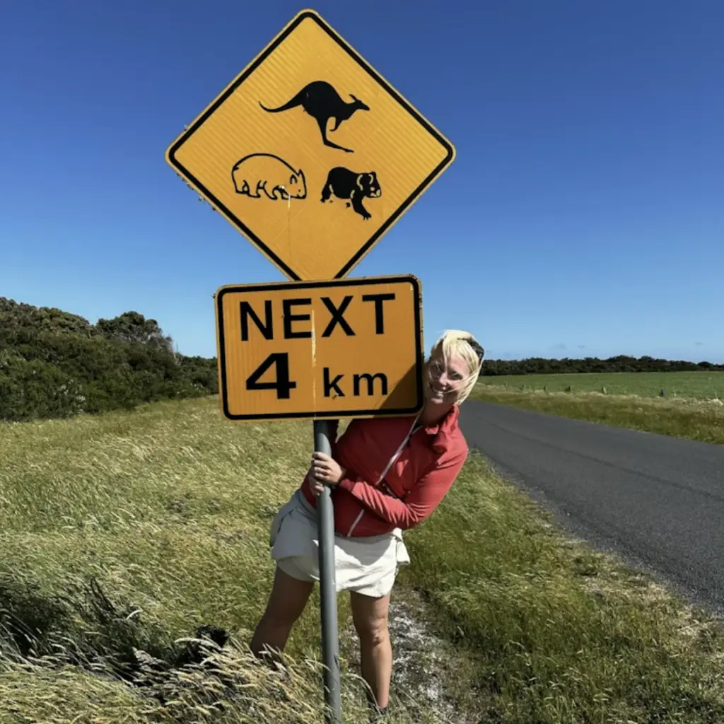 Frau steht neben einer Straße und umfasst ein australisches Verkehrsschild mit Tiersymbolen und Hinweis "Next 4 km"