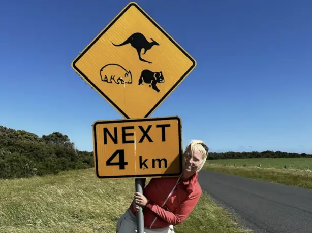 Frau steht neben einer Straße und umfasst ein australisches Verkehrsschild mit Tiersymbolen und Hinweis "Next 4 km"