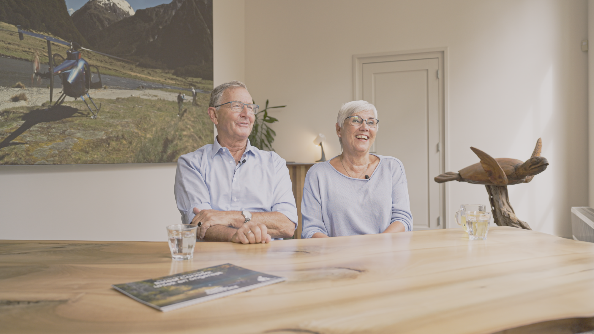 Klanten Job en Nelly aan een houten tafel tijdens een planningsgesprek met hun reisexpert, glimlachend terwijl ze hun aankomende reis bespreken, met een grote foto van een helikopterexcursie in Nieuw-Zeeland aan de muur achter hen.