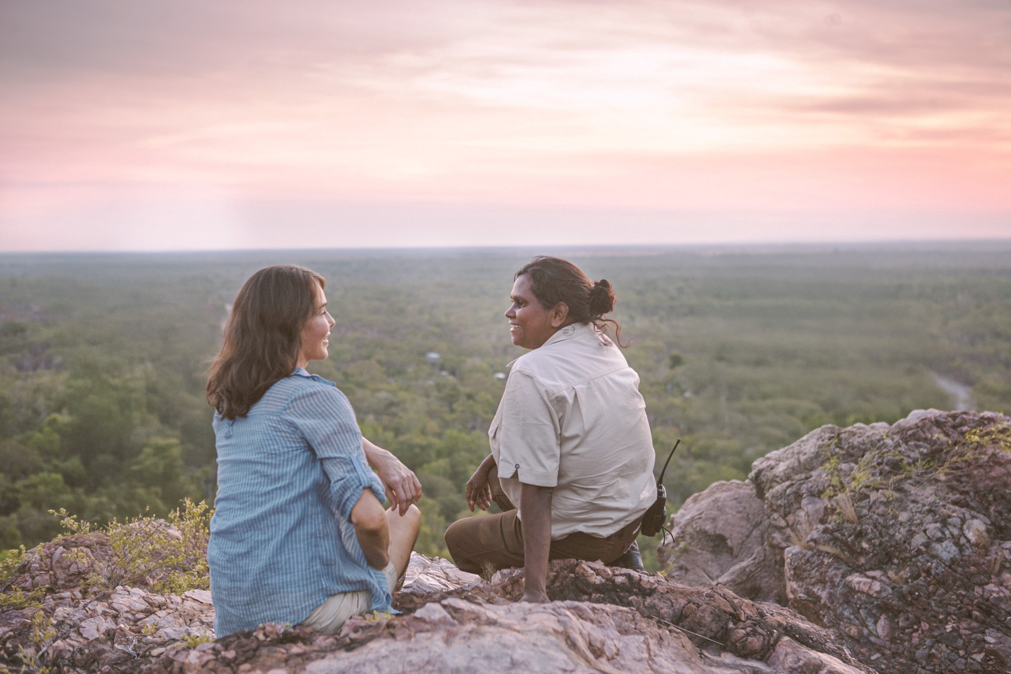 Een gids en een reiziger zitten op een rotsachtige steile helling met uitzicht op een uitgestrekt savannebos bij zonsondergang in Litchfield National Park, Northern Territory, Australië