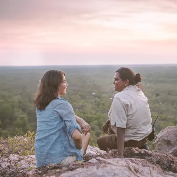 Ein Führer und Reisender sitzt bei Sonnenuntergang auf einer Felsklippe mit Blick auf ein weitläufiges Savannenwaldgebiet im Litchfield-Nationalpark, Northern Territory, Australien
