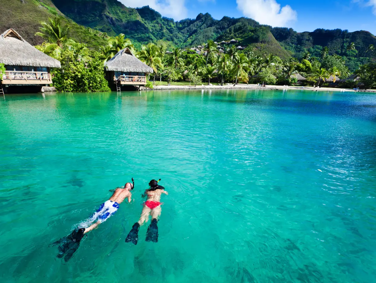 Twee mensen snorkelen in het turquoise water van Tahiti met waterhutten en bergen op de achtergrond