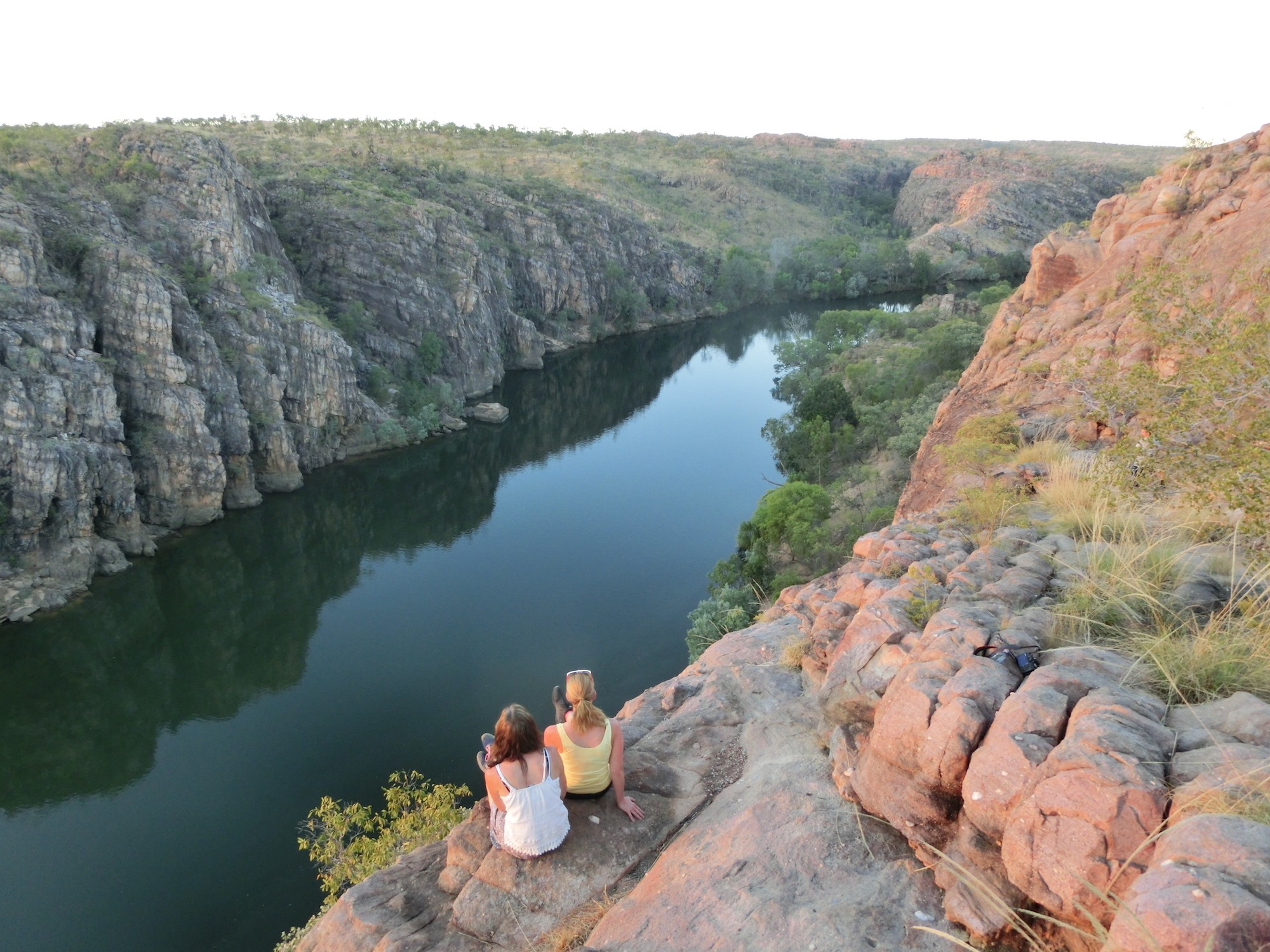 Two girls sitting at the edge of a cliff, looking out over a river