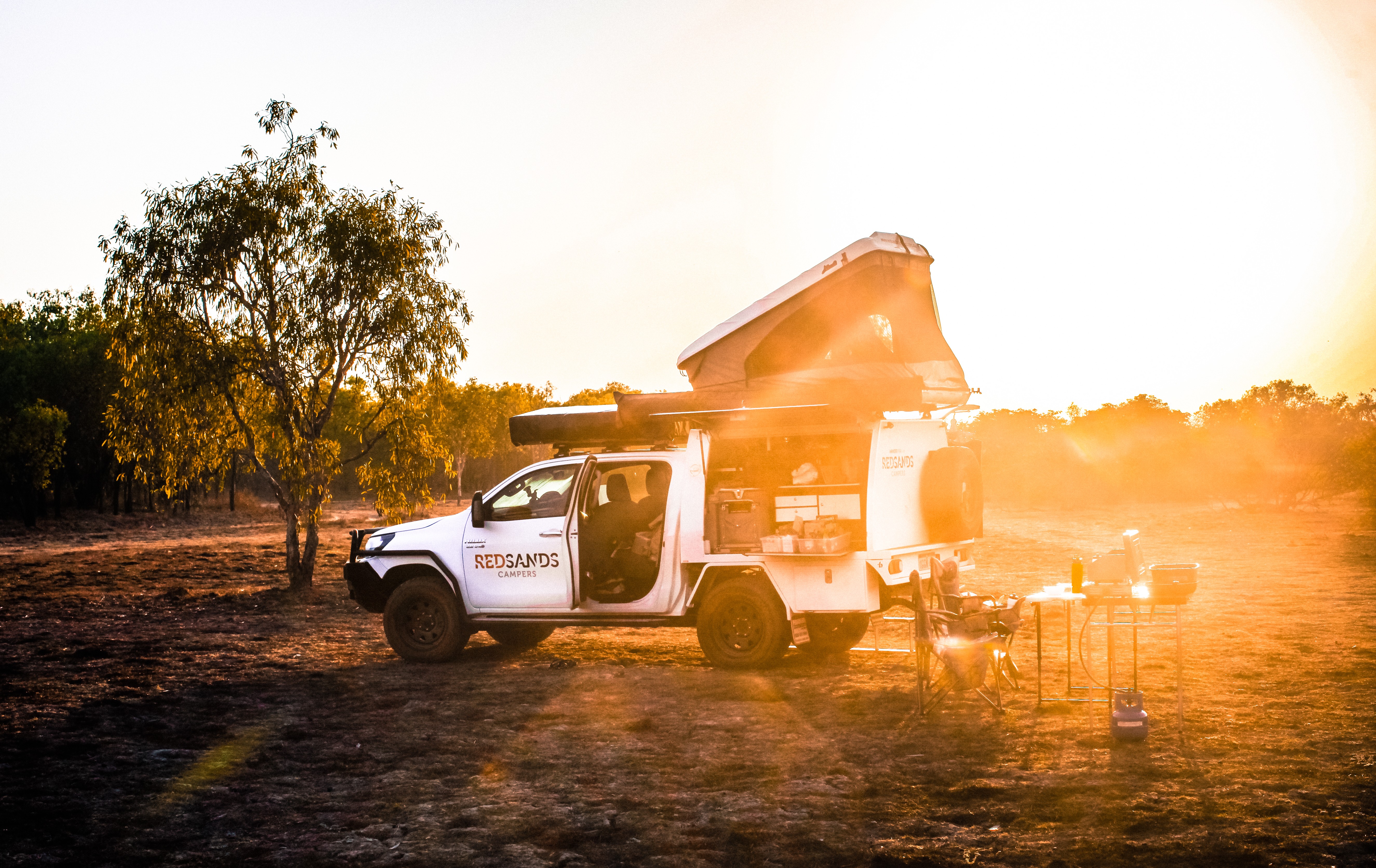 Ein Red Sands Campers 4WD mit Dachzelt und integrierter Campingküche auf einem Buschcampingplatz, getaucht in warmes goldenes Sonnenuntergangslicht zwischen Eukalyptusbäumen im australischen Outback, Australien.