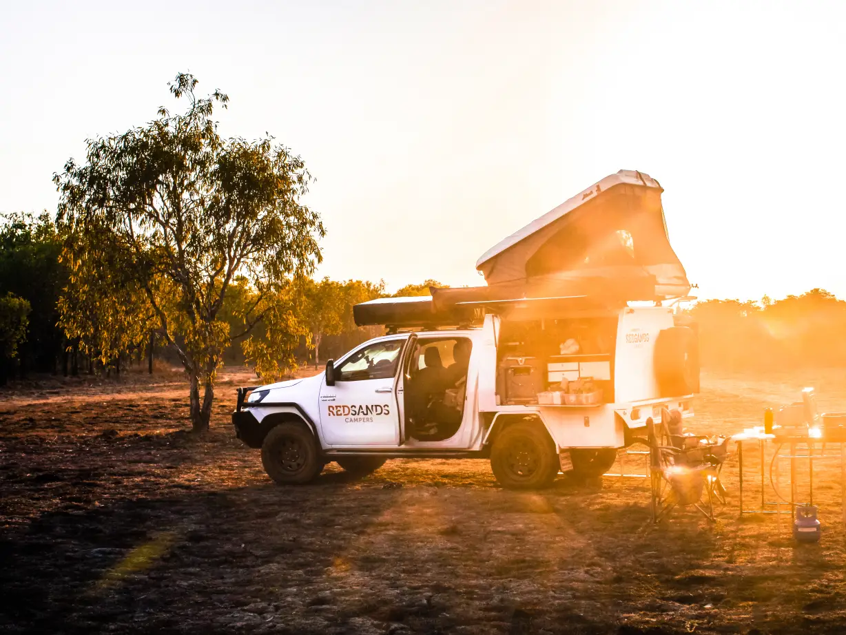 Ein Red Sands Campers 4WD mit Dachzelt und integrierter Campingküche auf einem Buschcampingplatz, getaucht in warmes goldenes Sonnenuntergangslicht zwischen Eukalyptusbäumen im australischen Outback, Australien.