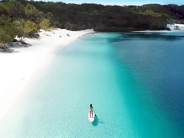 Ein Paddleboarder paddelt entlang eines weißen Sandstrandes auf K'Gari / Fraser Island, Queensland.