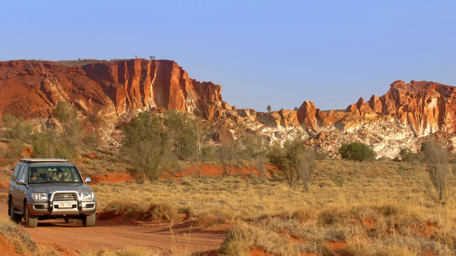 Off-road vehicle driving over red sand with red rocks in the background.