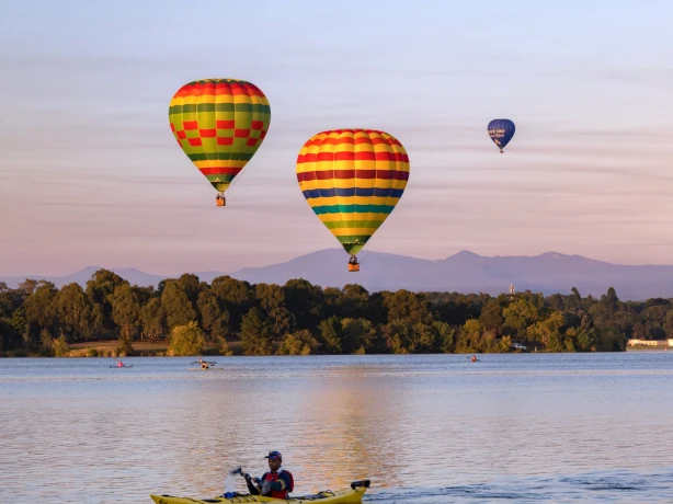 A kayaker paddles down a river in Canberra, Australia with hot air balloons flying overhead