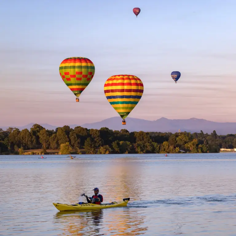 A kayaker paddles down a river in Canberra, Australia with hot air balloons flying overhead
