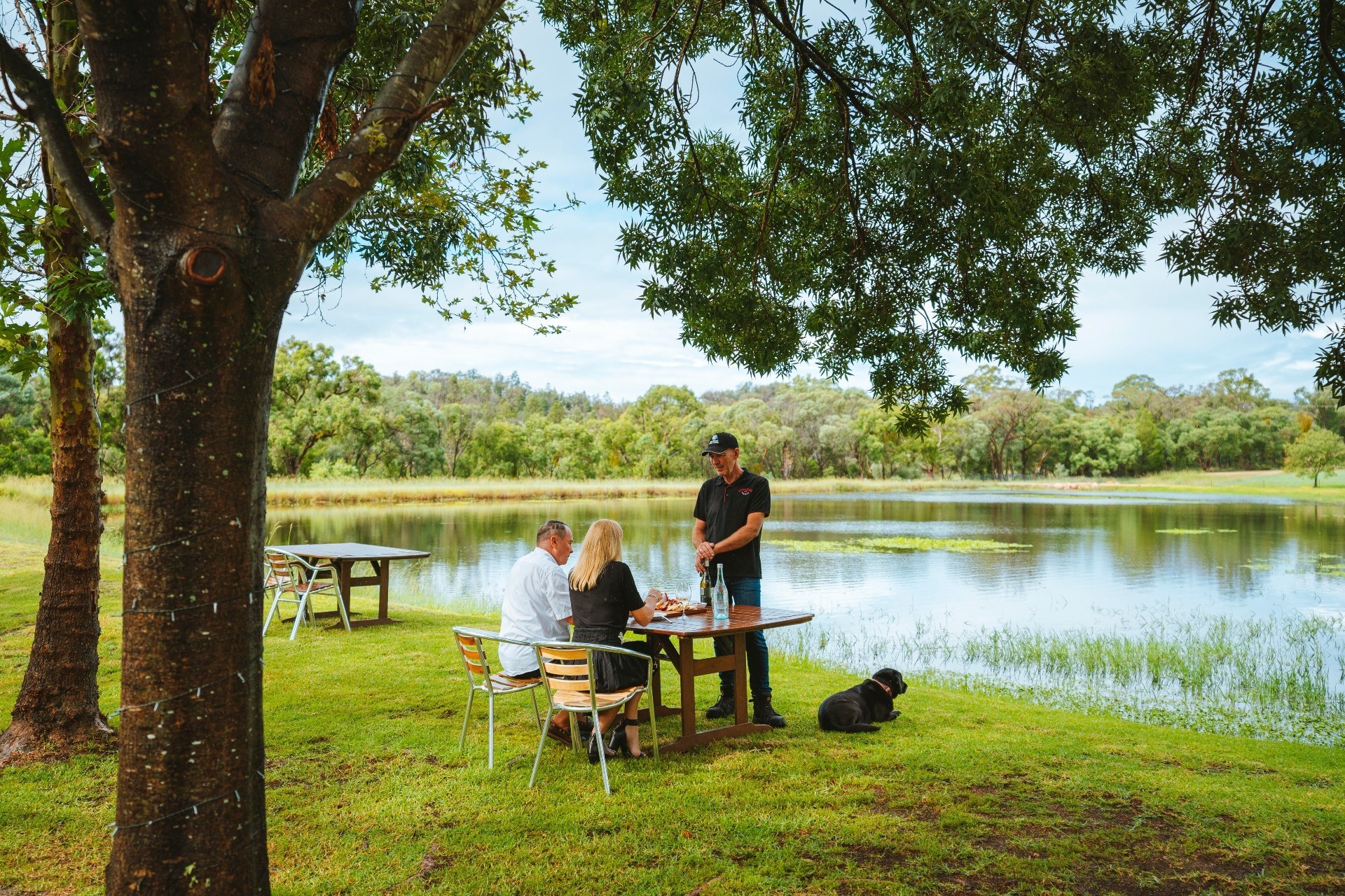 Ehepaar bei einer geführten Weinprobe an einem See mit einem schwarzen Hund in Australien
