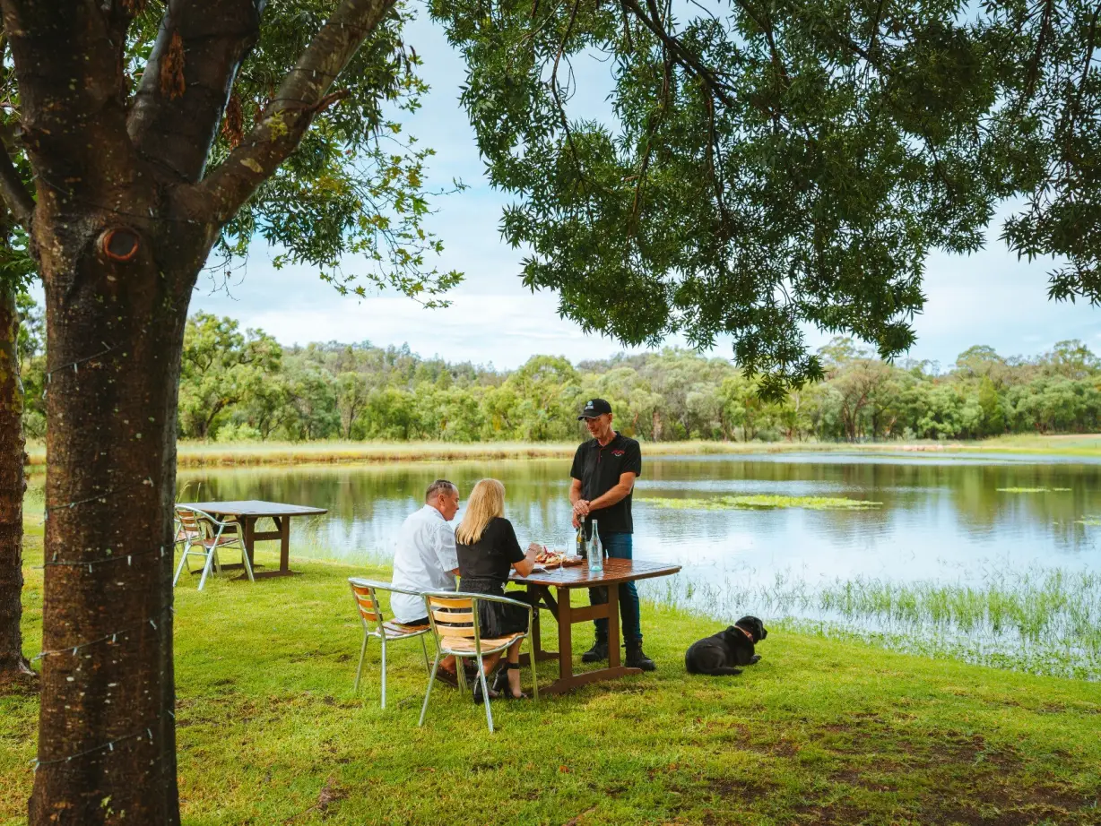 Ehepaar bei einer geführten Weinprobe an einem See mit einem schwarzen Hund in Australien