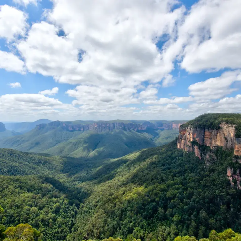 A landscape view of the Blue Mountains in New South Wales, Australia with abundant green trees and blue/cloudy sky