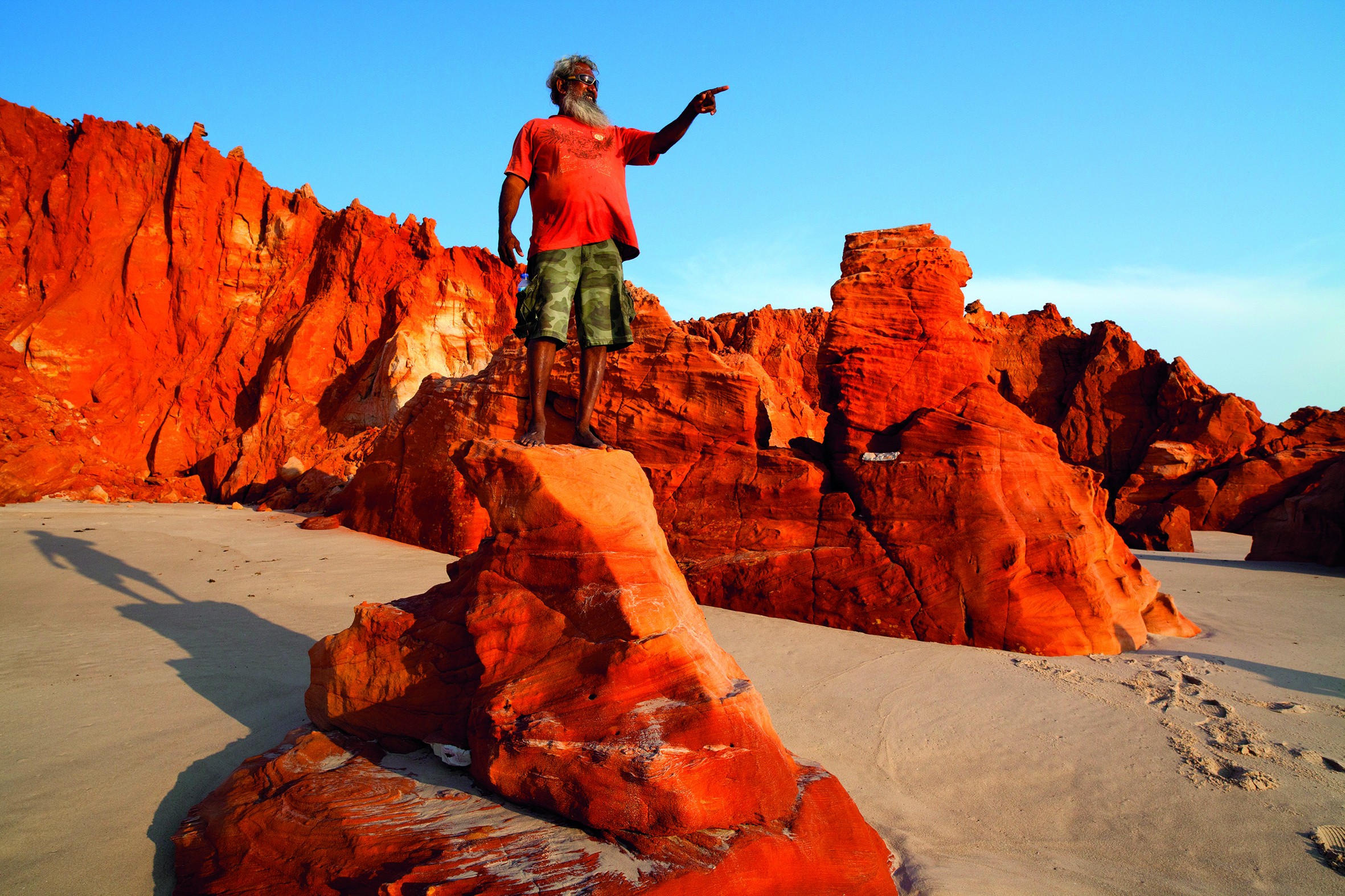 Ein Aborigine-Führer steht in Westaustralien auf einem rot gefärbten Felsen über hellem Sand und deutet auf etwas in der Ferne.