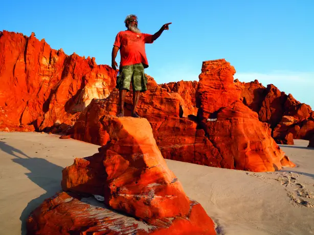 Ein Aborigine-Führer steht in Westaustralien auf einem rot gefärbten Felsen über hellem Sand und deutet auf etwas in der Ferne.