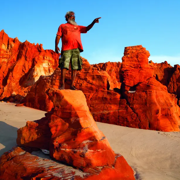 Ein Aborigine-Führer steht in Westaustralien auf einem rot gefärbten Felsen über hellem Sand und deutet auf etwas in der Ferne.