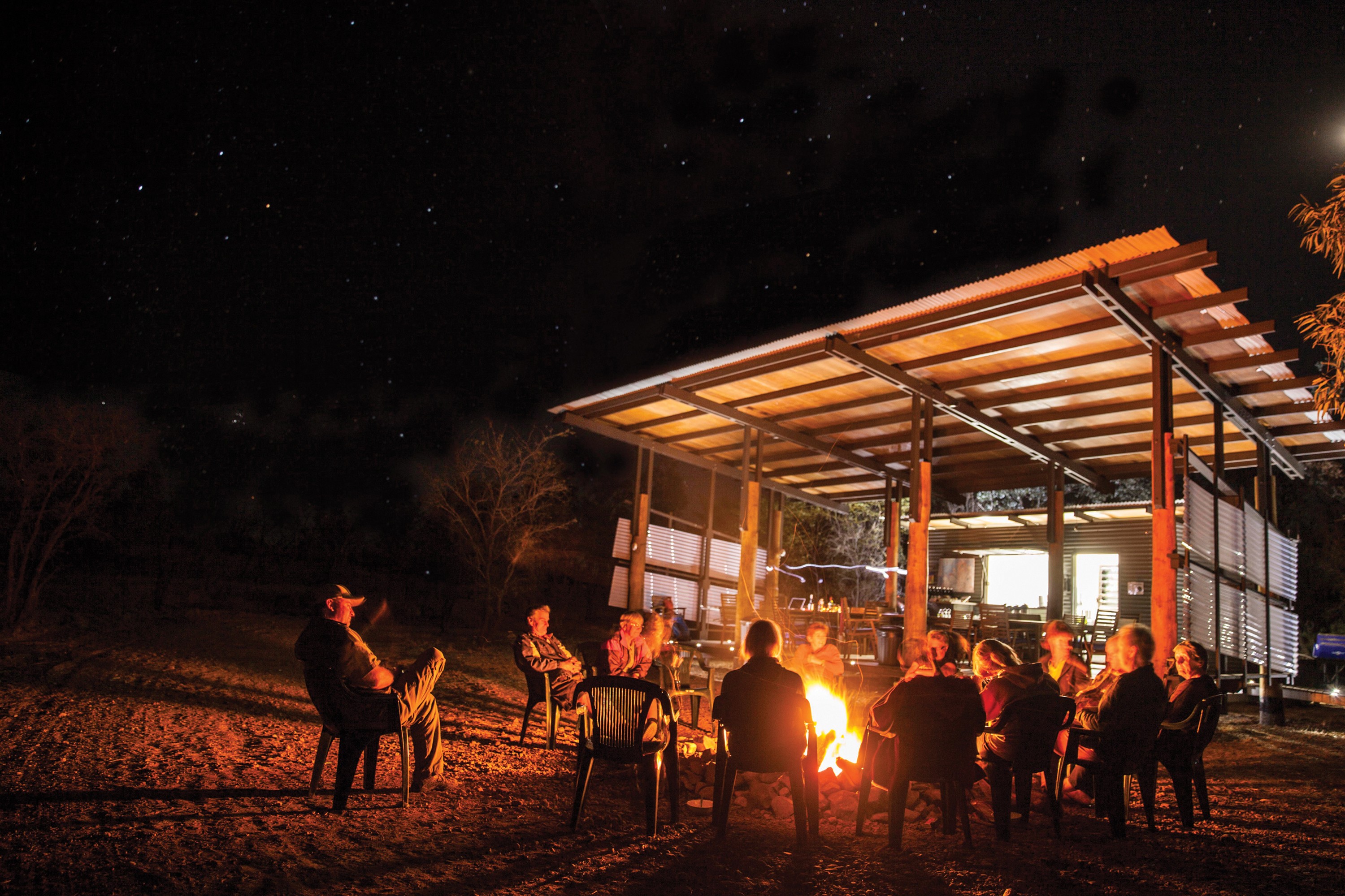 Mensen zitten bij een kampvuur onder een sterrenhemel naast een eenvoudig restaurantje van golfplaten in de Australische outback