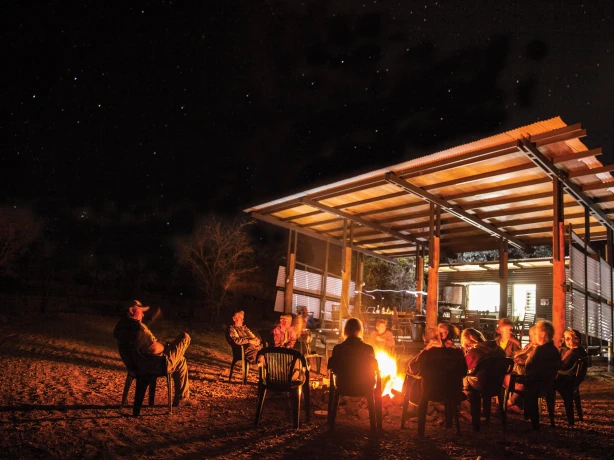 Mensen zitten bij een kampvuur onder een sterrenhemel naast een eenvoudig restaurantje van golfplaten in de Australische outback