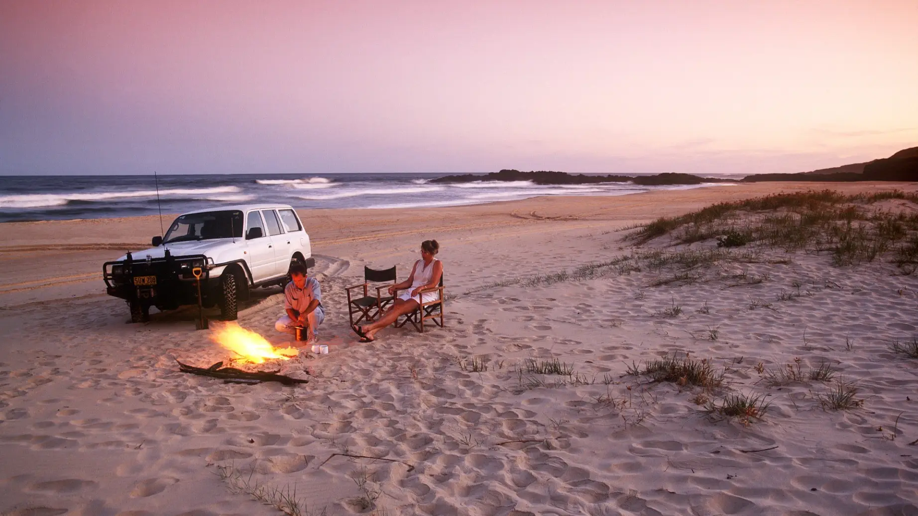 Two people sitting around a campfire on the beach next to a white car