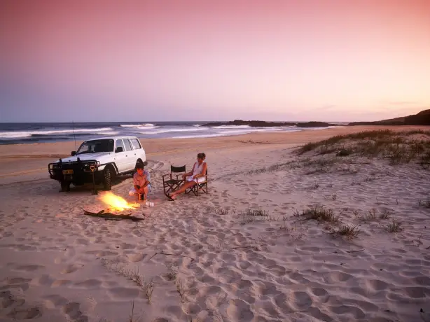Zwei Personen sitzen an einem Lagerfeuer am Strand neben einem weißen Auto