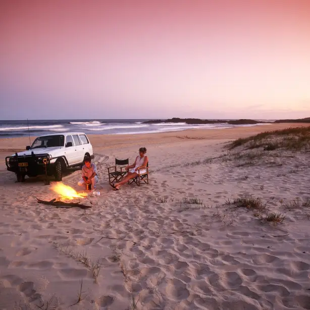 Zwei Personen sitzen an einem Lagerfeuer am Strand neben einem weißen Auto