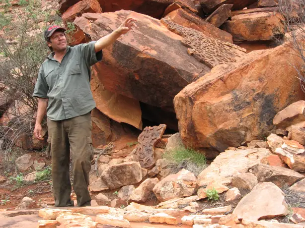 Ein indigener Führer zeigt auf uralte Felskunstgravuren auf großen roten Sandsteinbrocken während einer Kulturtour im australischen Outback, Australien.