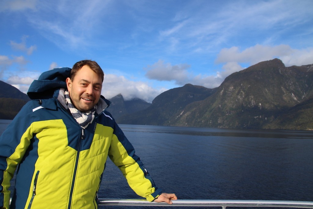 Mann steht an einer Reeling mit Gewässer und grünen Bergen im Hintergrund
