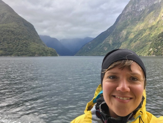 Frau mit Mütze und Regenmantel vor Wasser und Bergen des Doubtful Sound