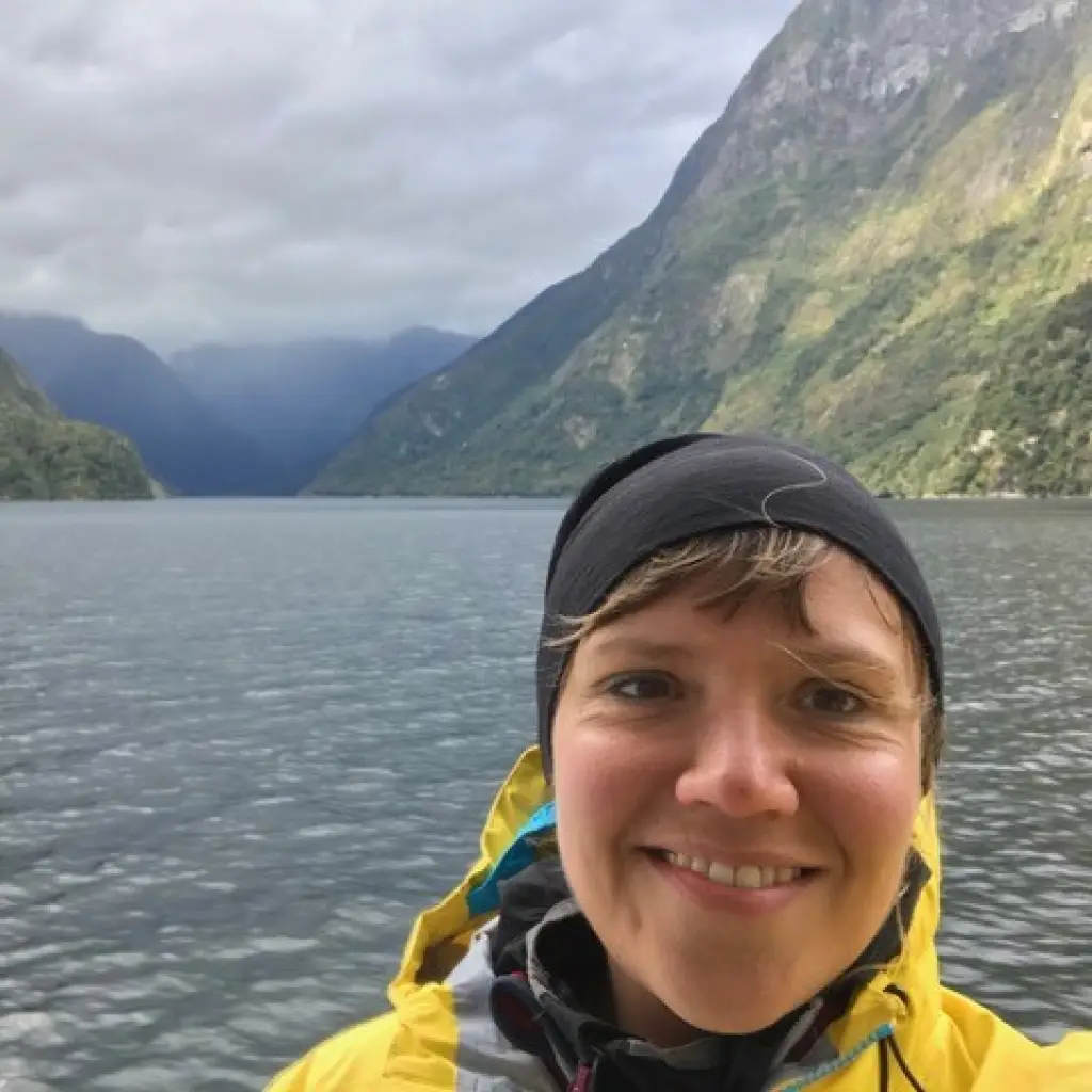 Frau mit Mütze und Regenmantel vor Wasser und Bergen des Doubtful Sound